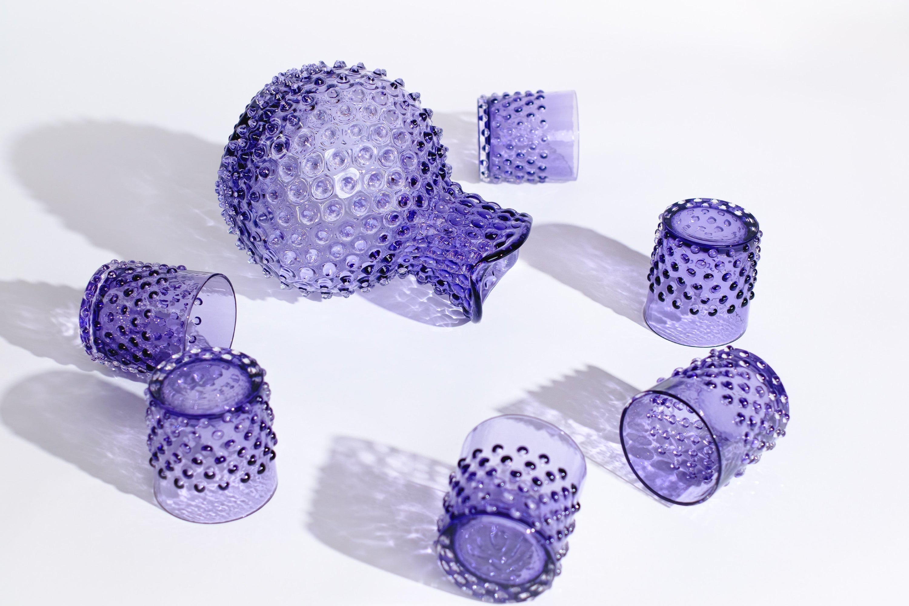Purple handmade glassware set with a dotted texture, including a pitcher and several tumblers, arranged on a white surface, casting shadows.